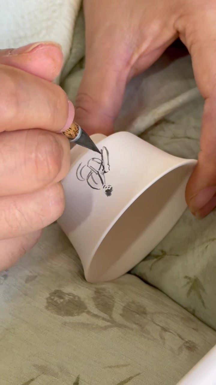 Tea Ware, Made by Hand A hand drawing dark lines on the side of a pale cup.